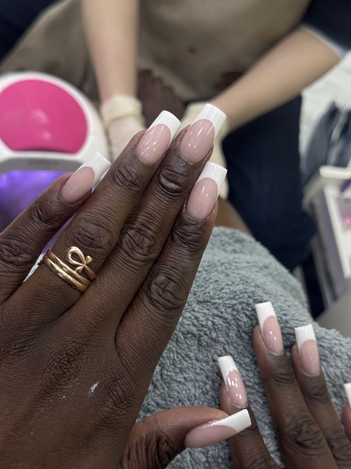 Two hands together showing long white French-tip nails, a technician working behind
